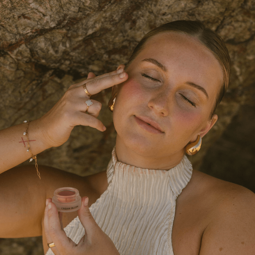 Woman applying Eco By Sonya Cream Blush to cheeks with finger, showing soft natural glow and smooth blendable texture.