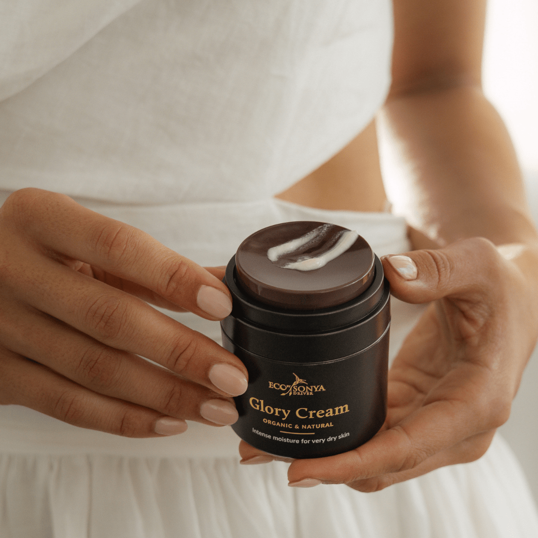 Hands holding Eco By Sonya Glory Cream jar with visible pump and creamy product smear, showing rich smooth texture against soft skin background.
