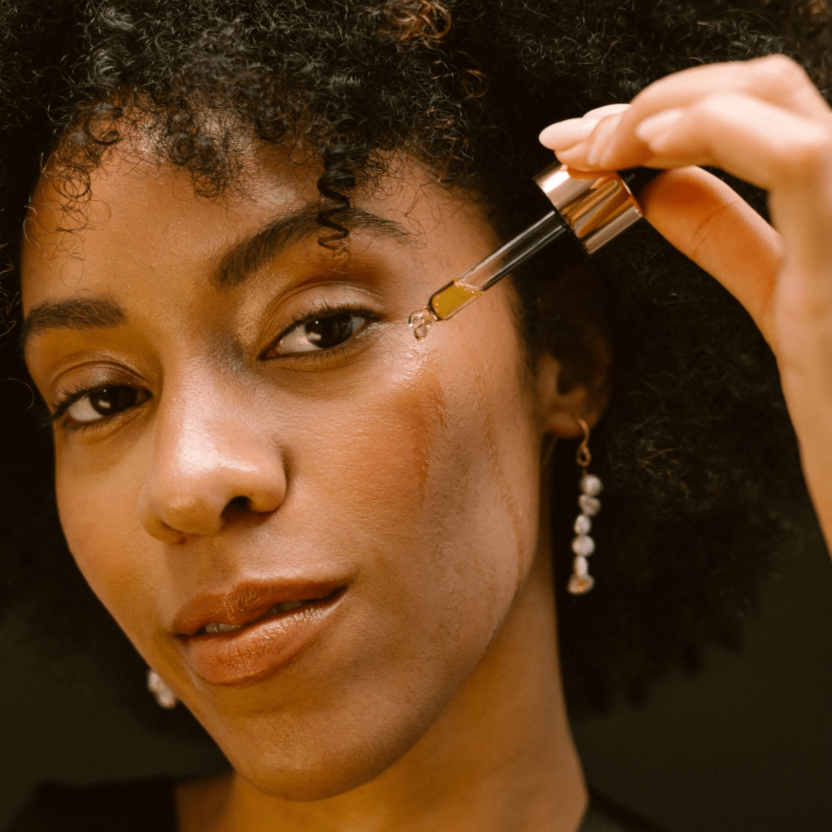 Woman applying a drop of Eco By Sonya Glory Oil onto her cheek, showing golden nourishing texture and hydrated radiant skin.