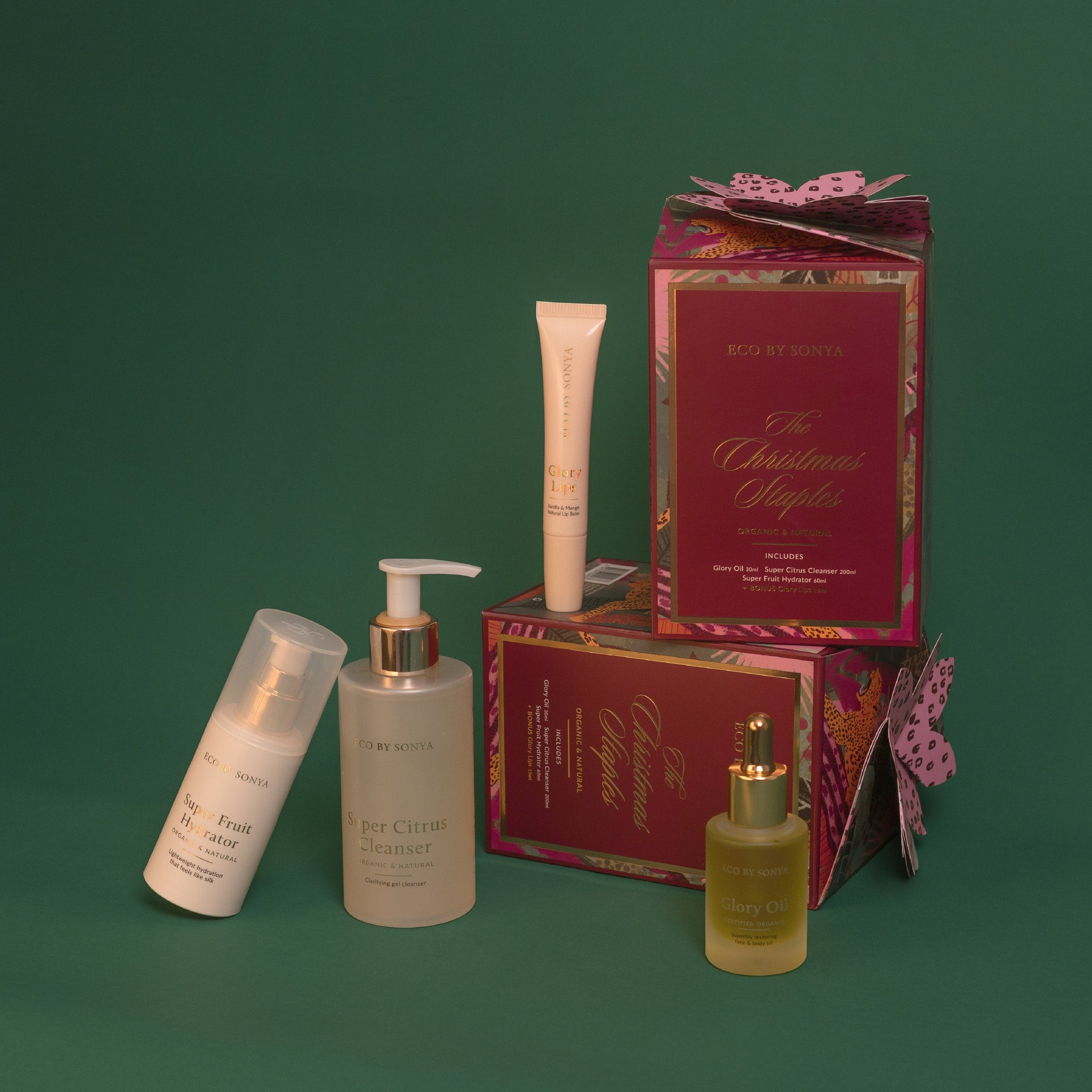 Eco By Sonya The Christmas Staples Gift Set featuring Super Citrus Cleanser, Super Fruit Hydrator, Glory Oil and bonus Glory Lips, arranged with decorative red and gold gift boxes on a dark green background.