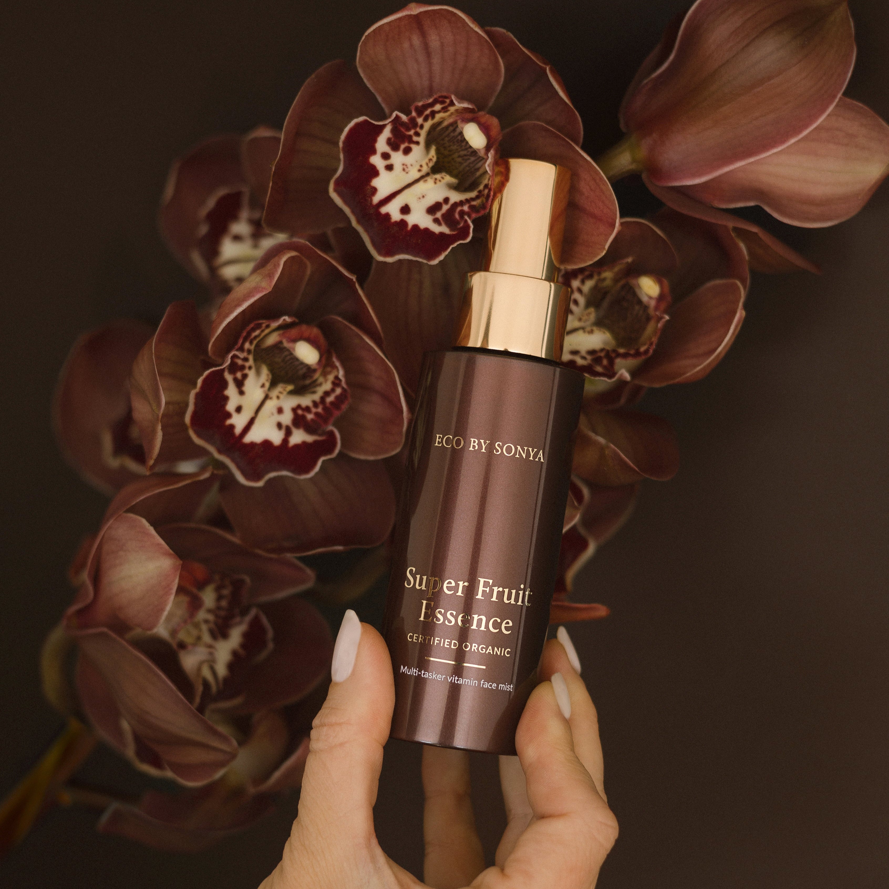 Hand holding a bottle of 'Super Fruit Essence' by Eco By Sonya against a dark background with flowers.