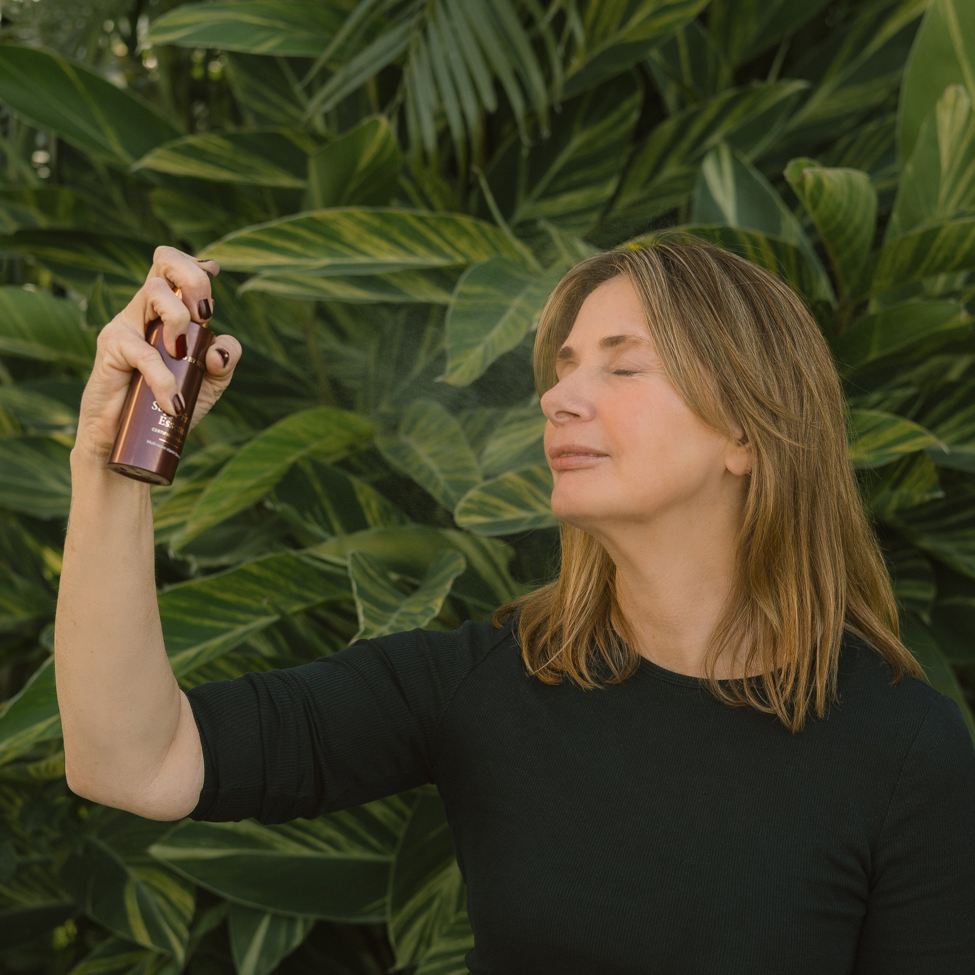 Woman spraying Eco By Sonya Super Fruit Essence onto her face, against a green leafy background