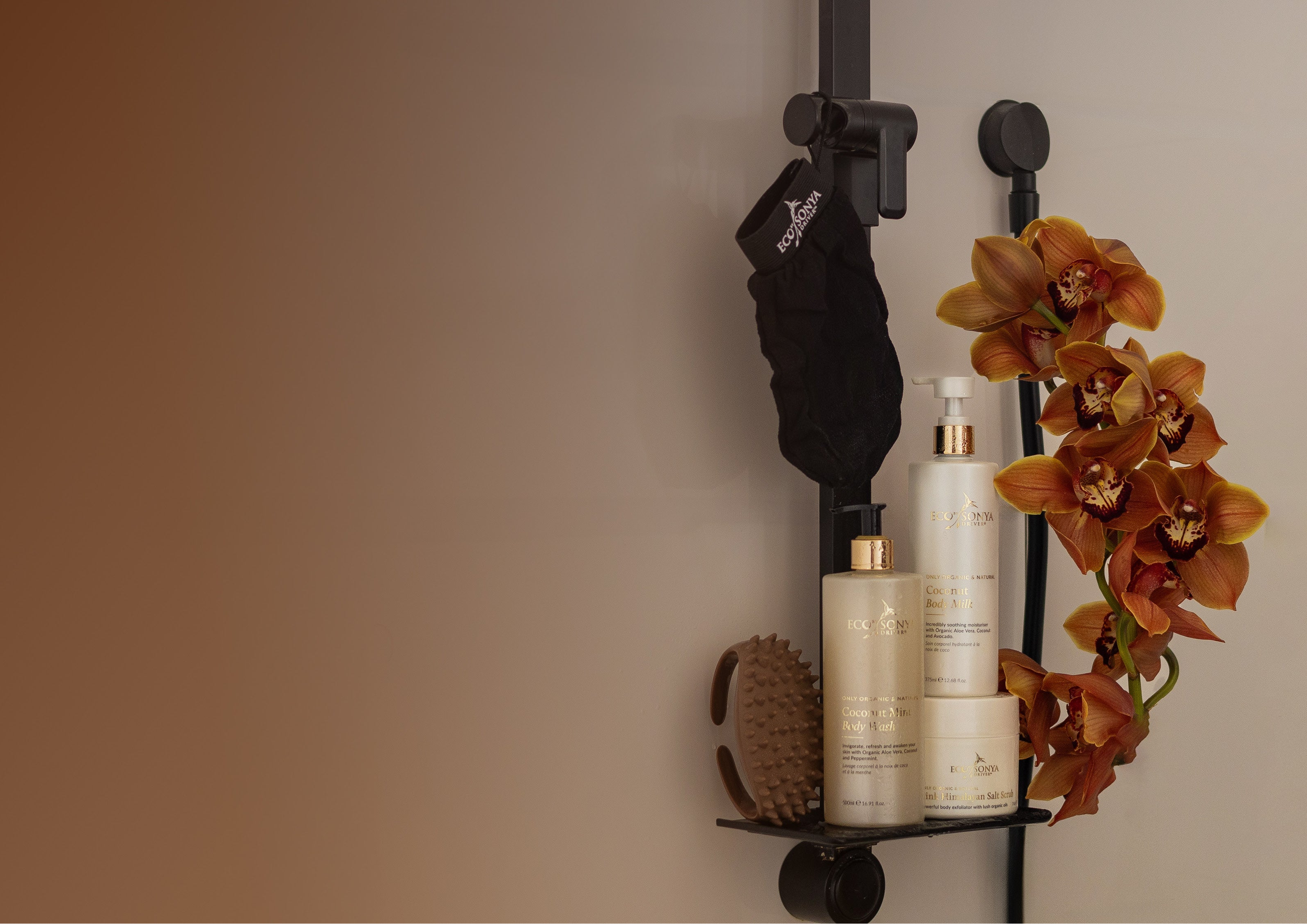 Bathroom shelf with Eco By Sonya Smooth Body Kit products and a flower arrangement against a beige wall.