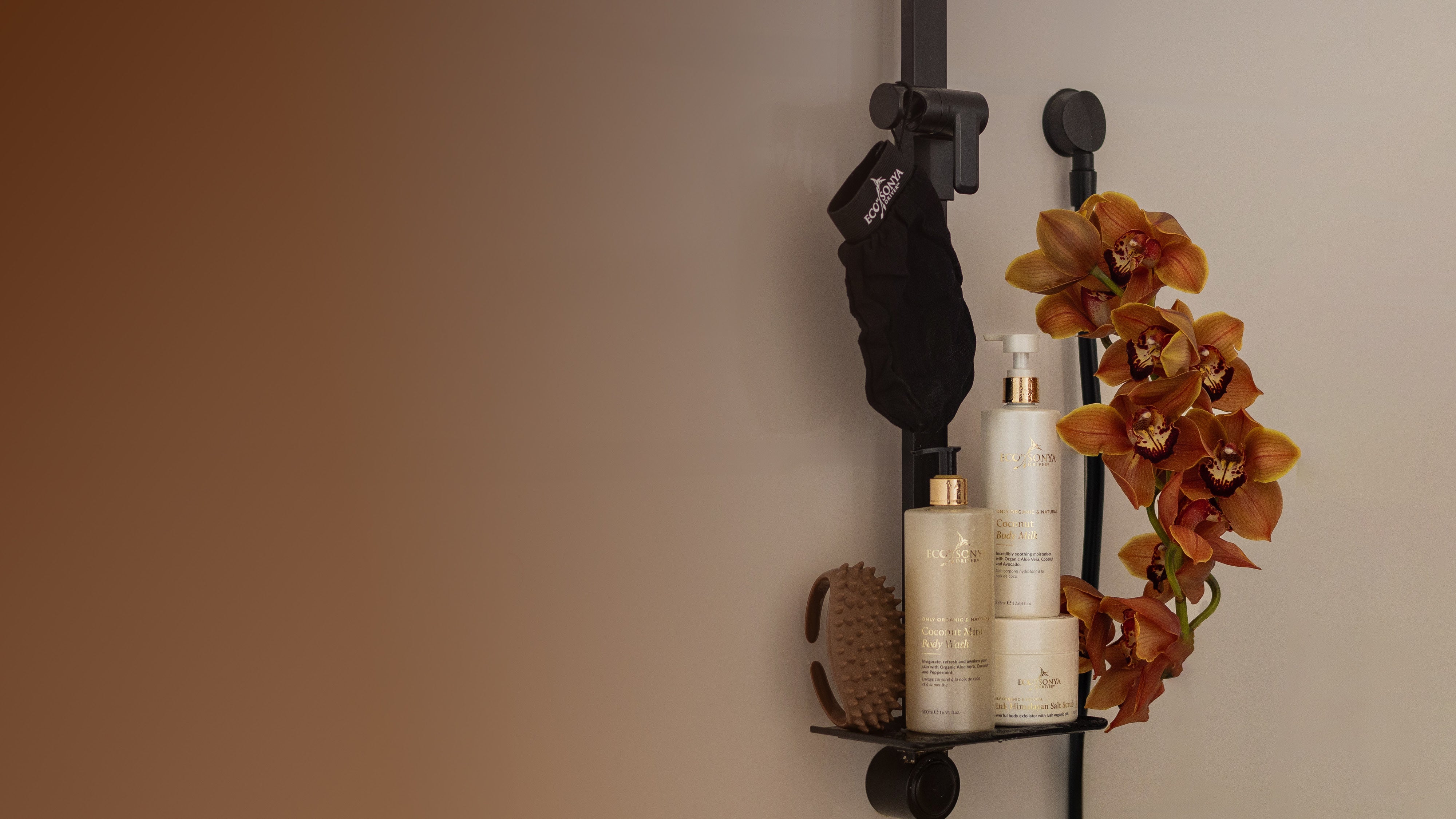 Bathroom shelf with Eco By Sonya Smooth Body Kit products and a flower arrangement against a beige wall.